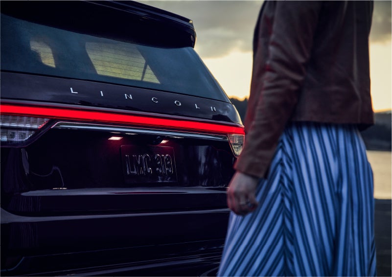 A person is shown near the rear of a 2023 Lincoln Aviator® SUV as the Lincoln Embrace illuminates the rear lights | Gettel Lincoln in Punta Gorda FL